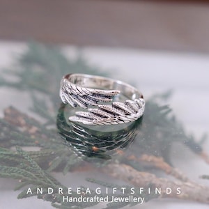 May include: A silver-colored ring with a feather design. The ring wraps around the finger with detailed feather textures. The ring is displayed on a reflective surface with the text "ANDREE A GIFTSFINDS Handcrafted Jewellery".