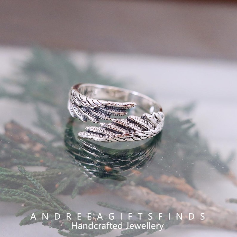 May include: A silver-colored ring with a feather design. The ring wraps around the finger with detailed feather textures. The ring is displayed on a reflective surface with the text "ANDREE A GIFTSFINDS Handcrafted Jewellery".