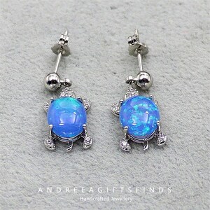 Könnte beinhalten: Silberne Ohrstecker mit Anhängern in Form von blauen Opal-Schildkröten. Jeder Ohrring hat ein Stecker- und Kugeldesign, wobei der Schildkrötenanhänger mit kleinen, klaren Edelsteinen verziert ist. Der Text "ANDREEA GIFTSFINDS Handcrafted Jewellery" befindet sich unten.