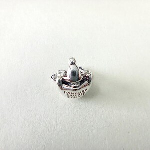 May include: A silver-colored Pandora charm with a rounded shape and a small, raised design on top. The charm has the Pandora logo and other text engraved on its surface. The charm is likely made of sterling silver.