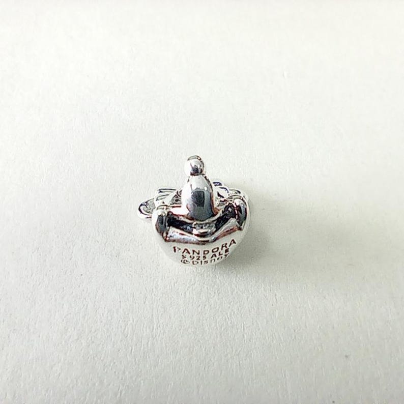 May include: A silver-colored Pandora charm with a rounded shape and a small, raised design on top. The charm has the Pandora logo and other text engraved on its surface. The charm is likely made of sterling silver.