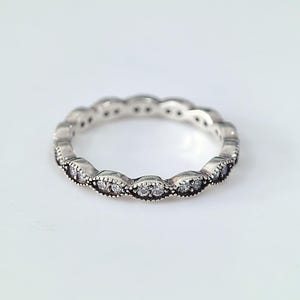Könnte beinhalten: Ein silberner Ring mit einem sich wiederholenden Muster aus ovalen Fassungen, die jeweils kleine, klare Edelsteine enthalten. Der Ring hat ein zartes, verziertes Design mit einer Vintage- oder Antik-Ästhetik. Die Edelsteine funkeln auf dem Silberband.