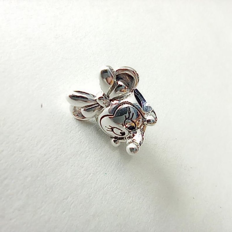 May include: A silver charm in the shape of a cartoon mouse with a bow. The charm has a detailed design, including the mouse's face, ears, and bow. The charm is made of a shiny, reflective metal.