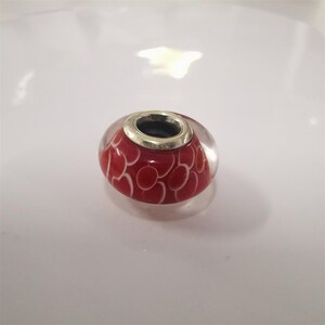 May include: A red and white glass bead with a silver-coloured metal rim. The bead features a pattern of white ovals on a red background. The bead is clear and transparent.