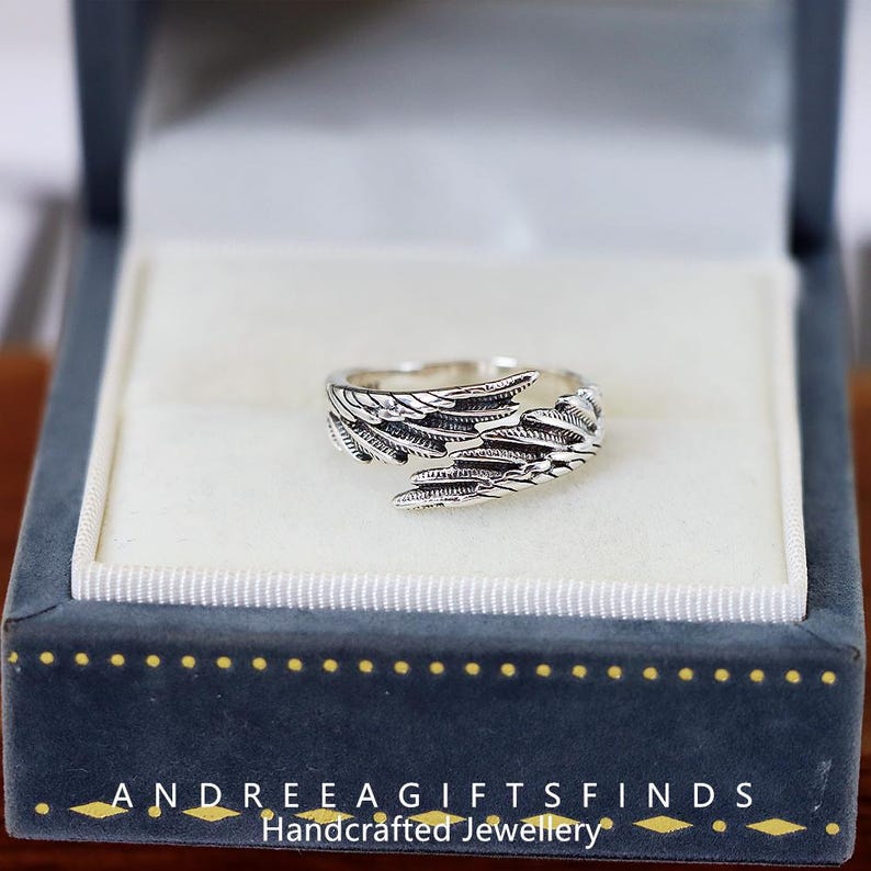 May include: A silver-colored ring with a wing design, displayed in a blue jewelry box. The ring features detailed feather-like patterns. The box has the text "ANDREEA GIFTSFINDS Handcrafted Jewellery".