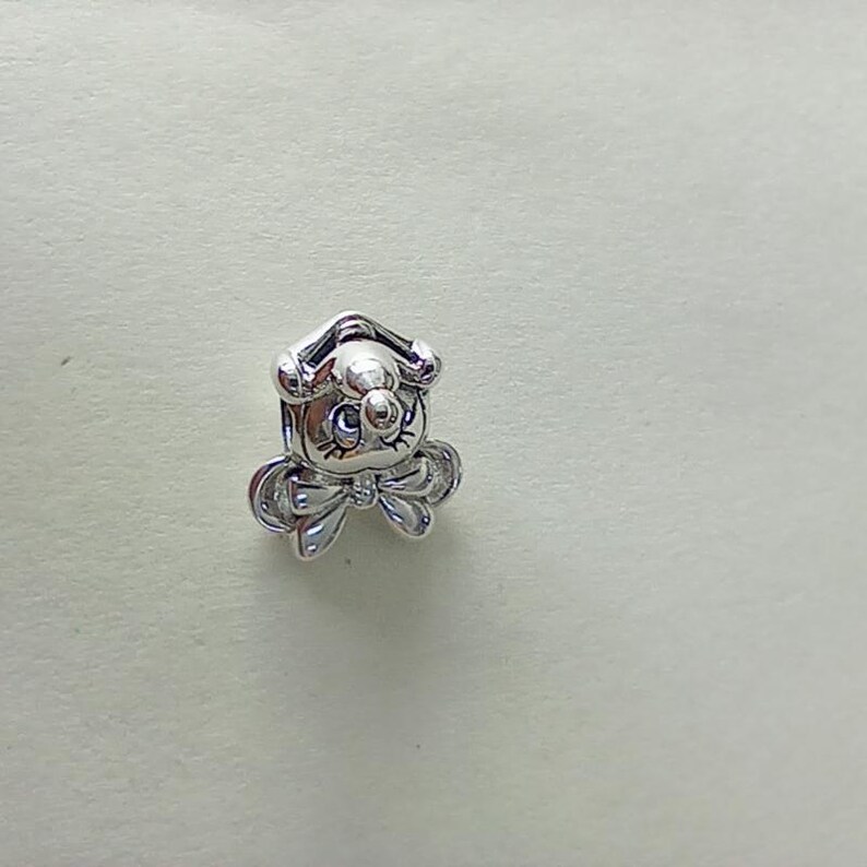 May include: A small, silver-colored charm in the shape of a cartoon dog with a bow tie. The charm has a detailed design, including the dog's face, ears, and bow. The charm is likely made of metal and is suitable for jewelry making or as a decorative element.