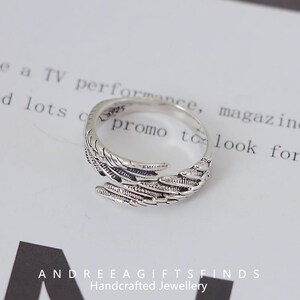 May include: A silver-colored ring with a feather design. The ring wraps around the finger with detailed feather textures. The ring is on a white surface with text in the background. The text "ANDREEAGIFTSFINDS Handcrafted Jewellery" is at the bottom.