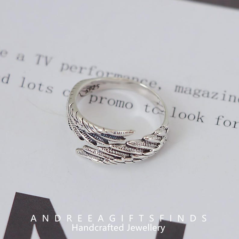 May include: A silver-colored ring with a feather design. The ring wraps around the finger with detailed feather textures. The ring is on a white surface with text in the background. The text "ANDREEAGIFTSFINDS Handcrafted Jewellery" is at the bottom.