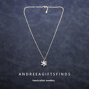 Könnte beinhalten: Eine zarte, goldfarbene Halskette mit einem Schneeflocken-Anhänger. Der Anhänger hat kleine, klare Edelsteine. Die Halskette wird auf einer dunkelblauen Oberfläche präsentiert. Der Text "ANDREEAGIFTSFINDS" und "Handcrafted Jewellery" befinden sich unten.