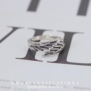 May include: A silver-colored ring with a detailed wing design. The ring features intricate feather detailing, creating a textured appearance. The ring is displayed on a white surface with text at the bottom that reads "ANDREEAGIFTSFINDS Handcrafted Jewellery."