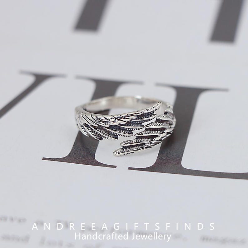 May include: A silver-colored ring with a detailed wing design. The ring features intricate feather detailing, creating a textured appearance. The ring is displayed on a white surface with text at the bottom that reads "ANDREEAGIFTSFINDS Handcrafted Jewellery."