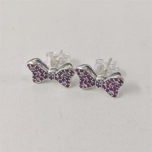 May include: A pair of silver bow-shaped stud earrings. Each bow is encrusted with small, round, dark pink gemstones. The earrings have a post and butterfly backing.