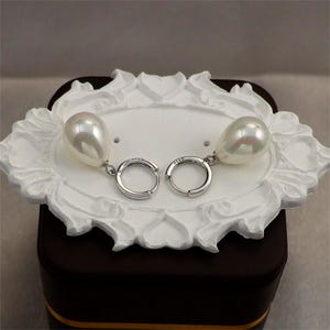 May include: A pair of pearl drop earrings with silver hoops. The earrings have large, teardrop-shaped pearls suspended from small, circular hoops. The earrings are displayed on a white, ornate decorative tray.