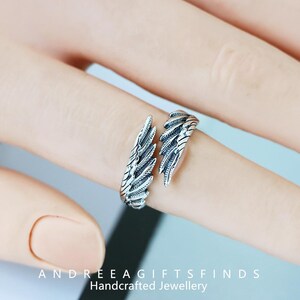 May include: A silver-colored ring in the shape of angel wings. The ring is open, with each wing wrapping around the finger. The ring is detailed with feather-like textures. The text "ANDREEA GIFTSFINDS Handcrafted Jewellery" is at the bottom.