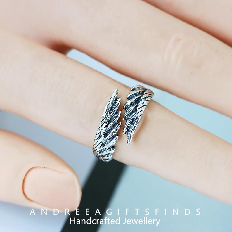 May include: A silver-colored ring in the shape of angel wings. The ring is open, with each wing wrapping around the finger. The ring is detailed with feather-like textures. The text "ANDREEA GIFTSFINDS Handcrafted Jewellery" is at the bottom.