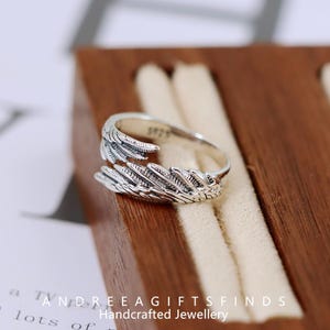 May include: A silver-colored ring with a feather design. The ring is detailed with individual feather barbs and is displayed on a wooden jewelry box. The text "ANDREEA GIFTSFINDS Handcrafted Jewellery" is visible.