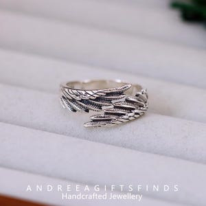 May include: A silver-colored ring with a detailed wing design. The ring features intricate feather detailing, with the wing wrapping around the finger. The text "ANDREEAGIFTSFINDS Handcrafted Jewellery" is at the bottom.