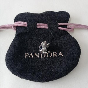 May include: A silver-colored charm in the shape of a cartoon character, resting on a black pouch with the brand name "PANDORA" printed in white. The pouch has a drawstring closure with a pink cord.