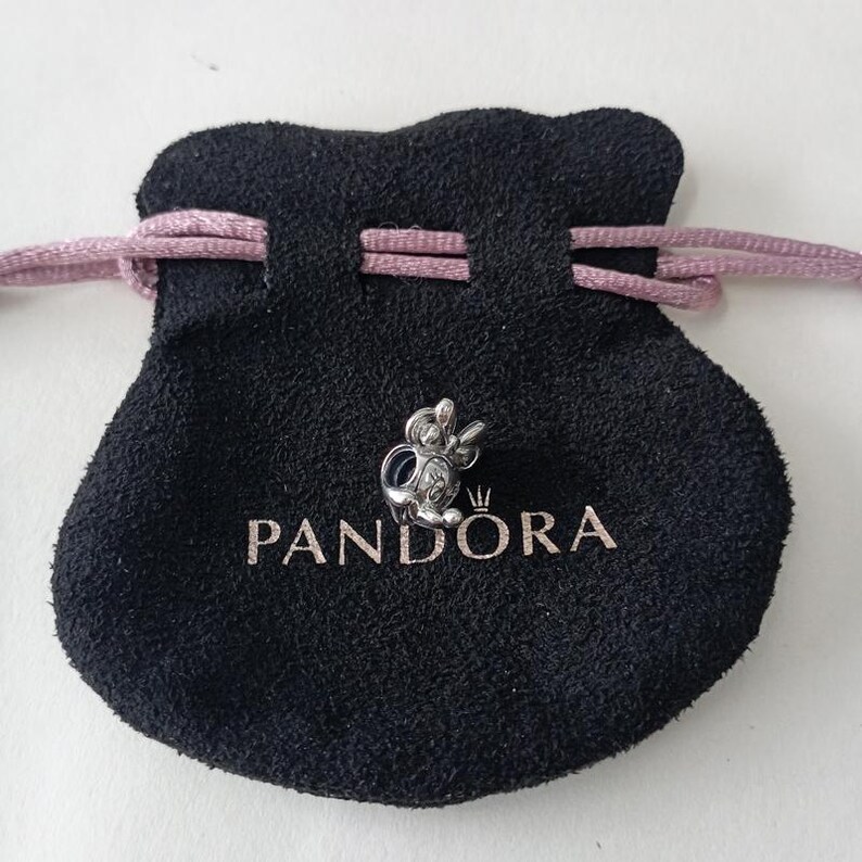 May include: A silver-colored charm in the shape of a cartoon character, resting on a black pouch with the brand name "PANDORA" printed in white. The pouch has a drawstring closure with a pink cord.