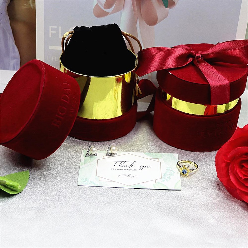 May include: A collection of jewelry gift boxes and accessories. The image features a red velvet jewelry box, a gold and red velvet jewelry box, a pair of pearl earrings, a gold ring with a blue stone, and a thank you card.