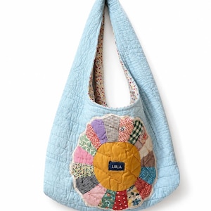 May include: A light blue quilted hobo bag featuring a large, circular, multi-colored patchwork flower appliqué on the front. The center of the flower is mustard yellow with a small navy blue rectangular tag reading LULA. The interior lining has a small floral print.