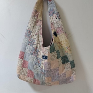 May include: A patchwork shoulder bag in pastel colors, including pink, blue, and cream. The bag is made of quilted fabric with a variety of floral and geometric patterns. A small, dark blue heart-shaped patch is sewn on the side, and a label reads "LULA".