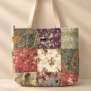 May include: A quilted tote bag featuring a patchwork design of various floral and paisley patterns in shades of pink, green, blue, and cream. The bag has two cream-colored fabric handles and a small black label with the text LULA.