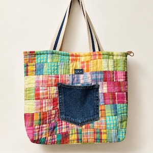 May include: A quilted tote bag made from a patchwork of multicolored plaid fabric squares. The bag features a dark blue denim pocket on the front, a small rectangular tag with the text LULA, and two beige and blue striped fabric handles.
