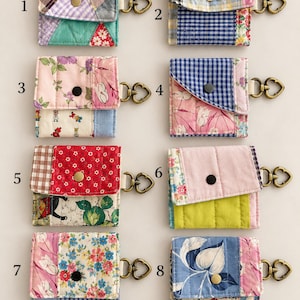 May include: Eight quilted fabric pouches labeled 1 through 8, each featuring a unique patchwork design, a snap button closure, and a bronze-toned heart-shaped metal clasp attached to the side. The pouches display various floral, plaid, and geometric patterns.