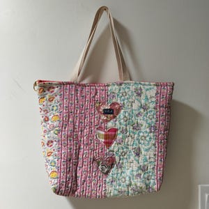 May include: A quilted tote bag with a patchwork design. The bag features a variety of floral and striped patterns in pink, white, and blue. Three heart-shaped appliqués are sewn onto the front. The bag has beige straps and the word "LULA" on one of the hearts.