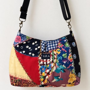 May include: A patchwork shoulder bag featuring various fabric patterns including floral, gingham, and polka dots in red, blue, yellow, and black. The bag has a black adjustable strap with metal hardware and a central label with the text LULA.