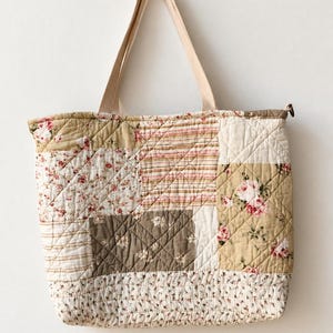 May include: A quilted tote bag featuring a patchwork design of various floral and striped fabric patterns in shades of beige, cream, pink, and brown. The bag has two light-colored fabric handles and a zippered top closure with a metal pull.