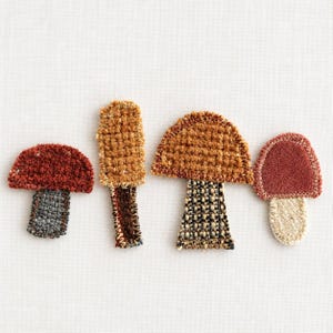 May include: Four embroidered mushroom patches arranged in a row. The first is dark red with a gray stem, the second is tan with a brown stem, the third is golden brown with a checkered stem, and the fourth is maroon with a cream stem.