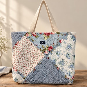 May include: A quilted tote bag featuring a patchwork design of floral patterns in red, blue, and white, alongside a navy and white gingham check fabric. The bag has two beige fabric handles and a small black rectangular label with the text LULA.
