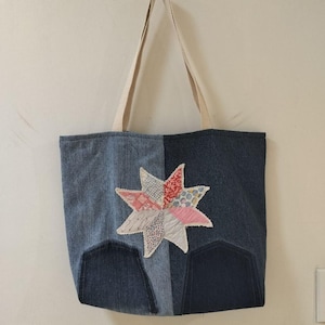 May include: Denim tote bag with a patchwork star design. The bag is made of two shades of blue denim and features a cream-colored handle. The star is made of various patterned fabrics in pink, red, and white.