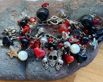 May include: A silver charm bracelet with a skull and crossbones charm, black and red heart charms, and other charms. The bracelet is on a gray rock background.