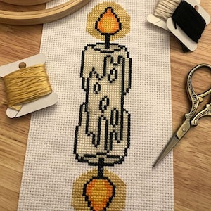 May include: Cross-stitch of a candle with a yellow flame and base, on white fabric. The candle is cream-colored with black detailing. Embroidery supplies, including thread spools and scissors, are arranged around the cross-stitch.
