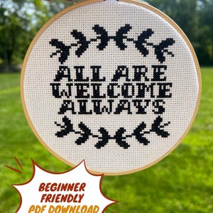 Puede incluir: Un bastidor de bordado de punto de cruz con las palabras "ALL ARE WELCOME ALWAYS" en hilo negro sobre tela blanca. El diseño incluye elementos florales negros decorativos. El texto "BEGINNER FRIENDLY PDF DOWNLOAD" está en un bocadillo rojo y amarillo.