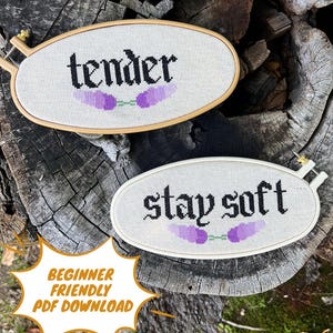 May include: Two oval cross-stitch projects in wooden hoops. One reads "tender" above a purple lavender sprig. The other says "stay soft" with a similar lavender design. A white speech bubble reads "BEGINNER FRIENDLY PDF DOWNLOAD".