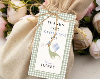 Peut inclure: Un sac cadeau beige avec cordon de serrage et une étiquette avec l'inscription "THANKS FOR STOMPING BY!" et "love, HENRY". L'étiquette représente un dinosaure vert avec des ballons. L'étiquette a une bordure à carreaux verts et blancs et est attachée avec de la ficelle.