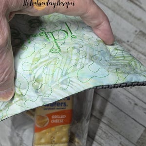 May include: A clear glass jar filled with a package of grilled cheese crackers, topped with a quilted lid. The lid features a green embroidered monogram and a light green and blue pattern. The jar is held by a hand.