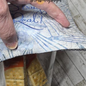 May include: A clear glass jar filled with packaged crackers, partially covered by a quilted fabric lid. The lid is white and blue with embroidered text that reads "Real!" The jar is held by a hand.