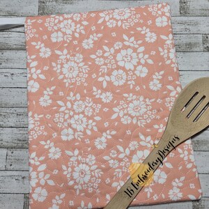 May include: A rectangular quilted potholder in a soft peach color featuring a white floral pattern and a white hanging loop, paired with a wooden slotted spoon that has the text IB Embroidery Designs printed on the handle.