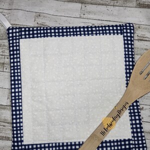 Può includere: Un sottopentola imbottito bianco con un bordo a quadri blu e bianchi. Il sottopentola ha un anello per appenderlo. Un cucchiaio di legno è appoggiato sul sottopentola.