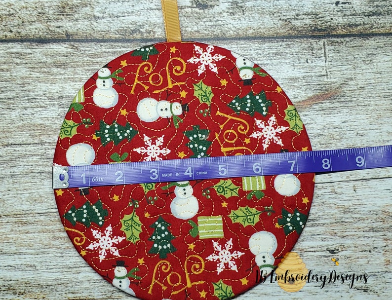 K&ouml;nnte beinhalten: Ein runder, roter Weihnachtsschmuck mit einem festlichen Muster mit Schneem&auml;nnern, Schneeflocken und Stechpalmen. Der Schmuck hat einen Durchmesser von etwa 18 cm und ein goldenes Band zum Aufh&auml;ngen. Das Design beinhaltet das Wort "JOY".