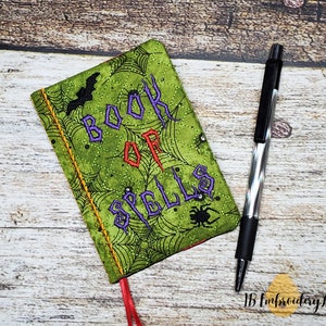 May include: A green fabric book cover with a spiderweb pattern and the text "BOOK OF SPELLS" in purple and red lettering. A black pen is lying next to the book.