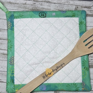 Könnte beinhalten: Ein grüner und blauer gesteppter Topflappen mit einem Blumenmuster. Ein Holzlöffel mit dem Text "LB Embroidery Designs" liegt auf dem Topflappen.