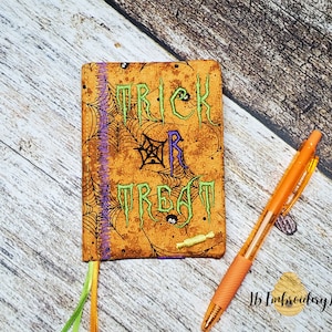 May include: A small orange and brown fabric book with the words "Trick or Treat" embroidered on the front. The book has a purple and green ribbon attached to the spine. A pen is lying on a wooden surface next to the book.