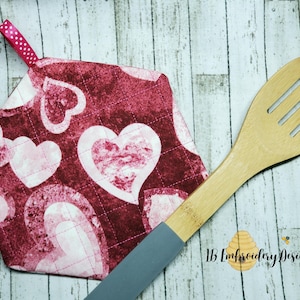 May include: Red and white quilted hot pad with a heart pattern. The hot pad has a pink ribbon loop for hanging.  The hot pad is next to a wooden slotted spoon.