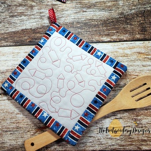 May include: A white pot holder with a red stitched design of fireworks and a blue and red striped border. The pot holder is on a wooden surface with a wooden spoon.